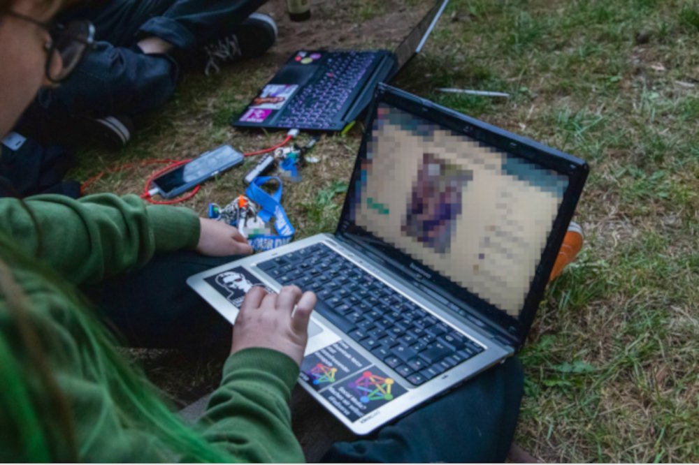 Ich von schräg hinten, wie ich einen Laptop auf dem Schoß habe. Der Bildschirm ist verpixelt, im Hintergrund sind andere Menschen mit Laptops auf einer Wiese zu erkennen Ückück Stephanie Henkel Laptop